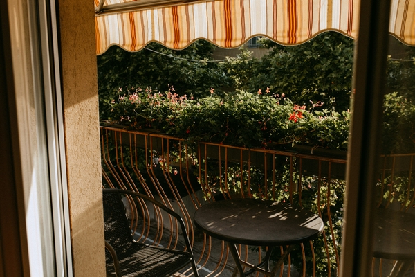 Ein schattiger Balkon mit farbenfrohen Blumen und einem runden Tisch, gesehen durch eine geöffnete Balkontür.